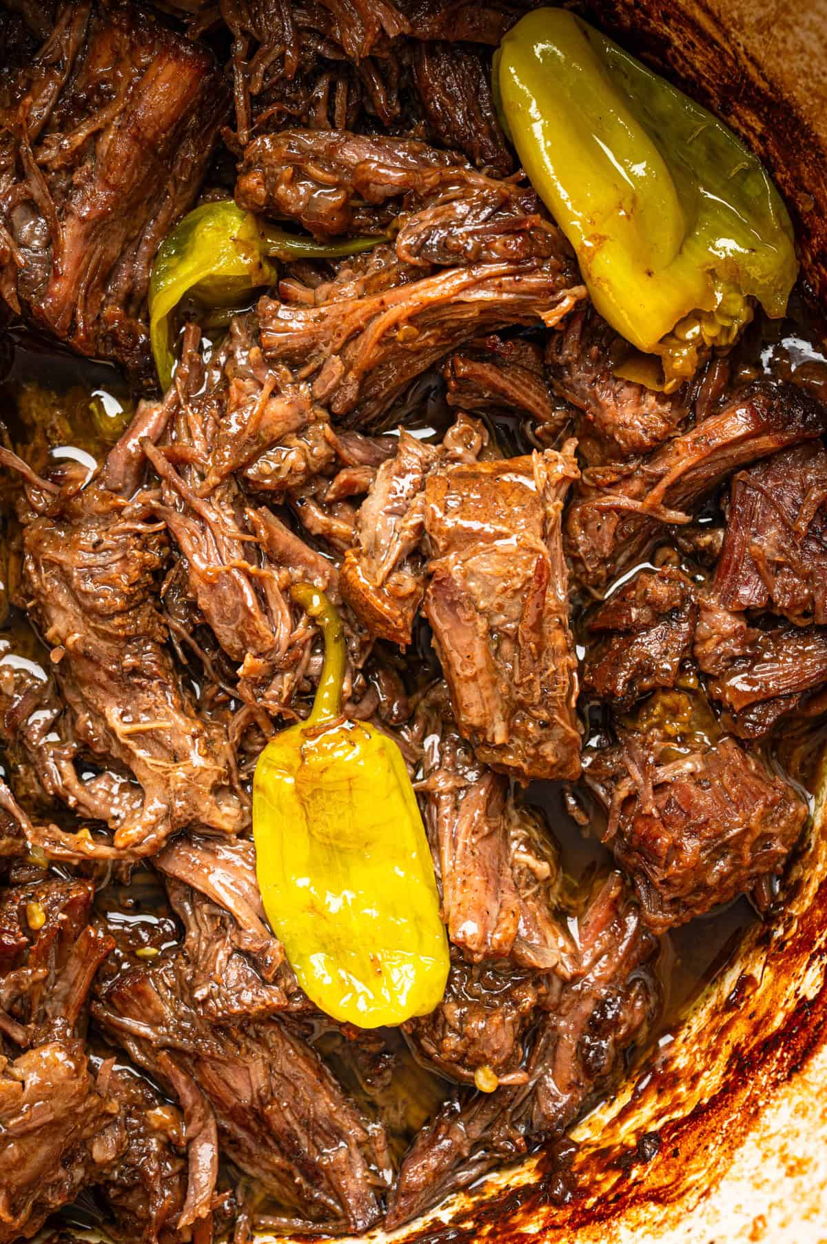 Mississippi Pot Roast (Easy Oven-Braised Chuck Roast) Shredded, juicy beef cooked with yellow pepperoncini peppers in a rich, savory sauce, shown in a close-up view in a pot.