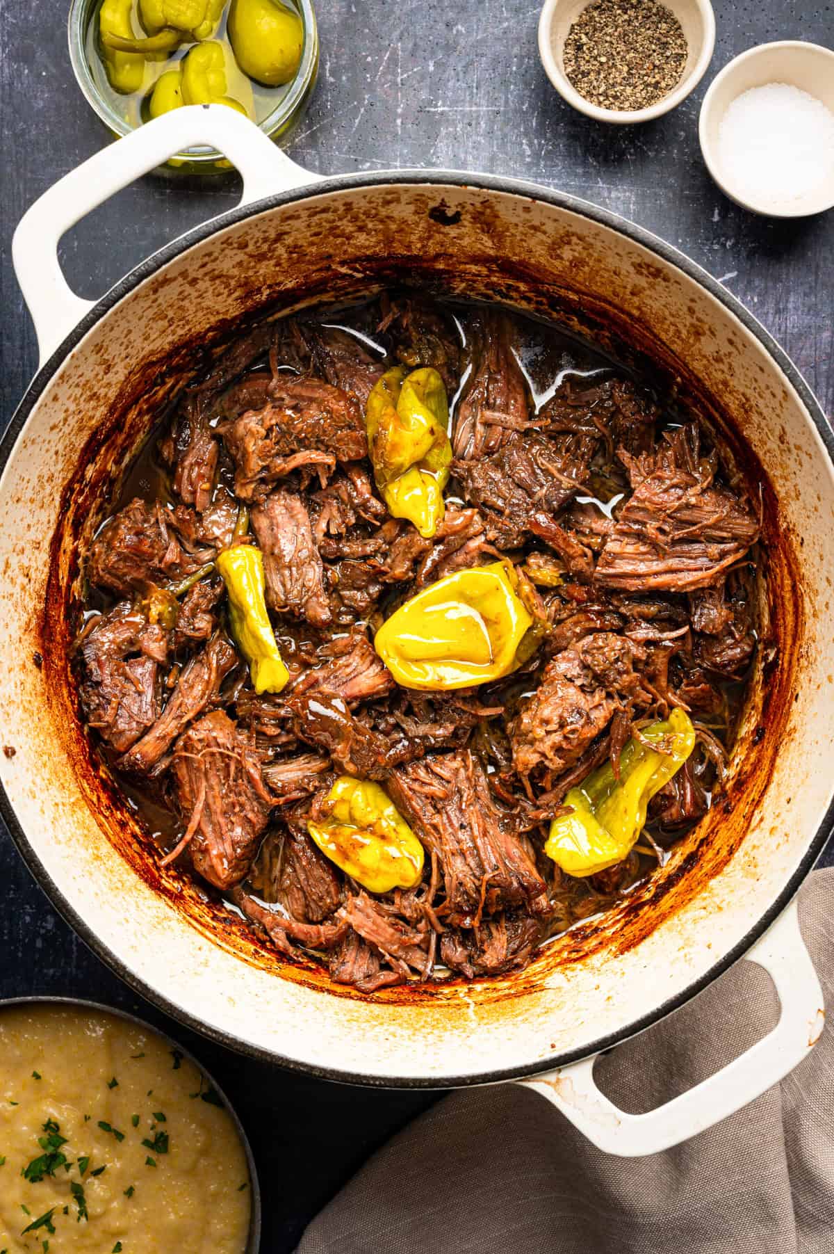 Mississippi Pot Roast (Easy Oven-Braised Chuck Roast) Shredded pot roast with several yellow pepperoncini peppers in a Dutch oven.