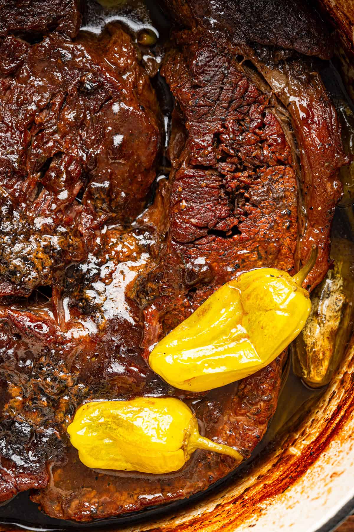 Mississippi Pot Roast (Easy Oven-Braised Chuck Roast) Close-up of cooked pot roast with two yellow pepperoncini peppers on top, surrounded by a rich, dark broth in a pot.