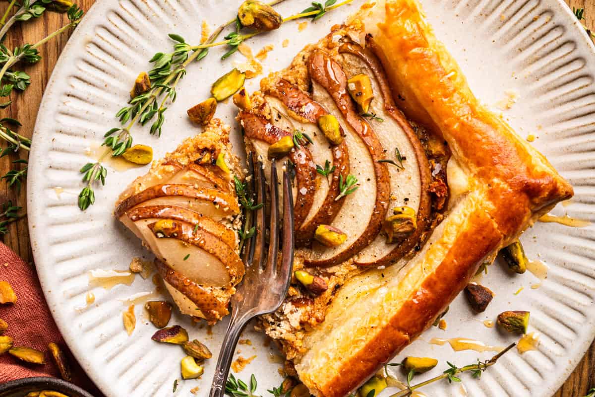 A slice of pear tart with pistachios and herbs on a plate. 