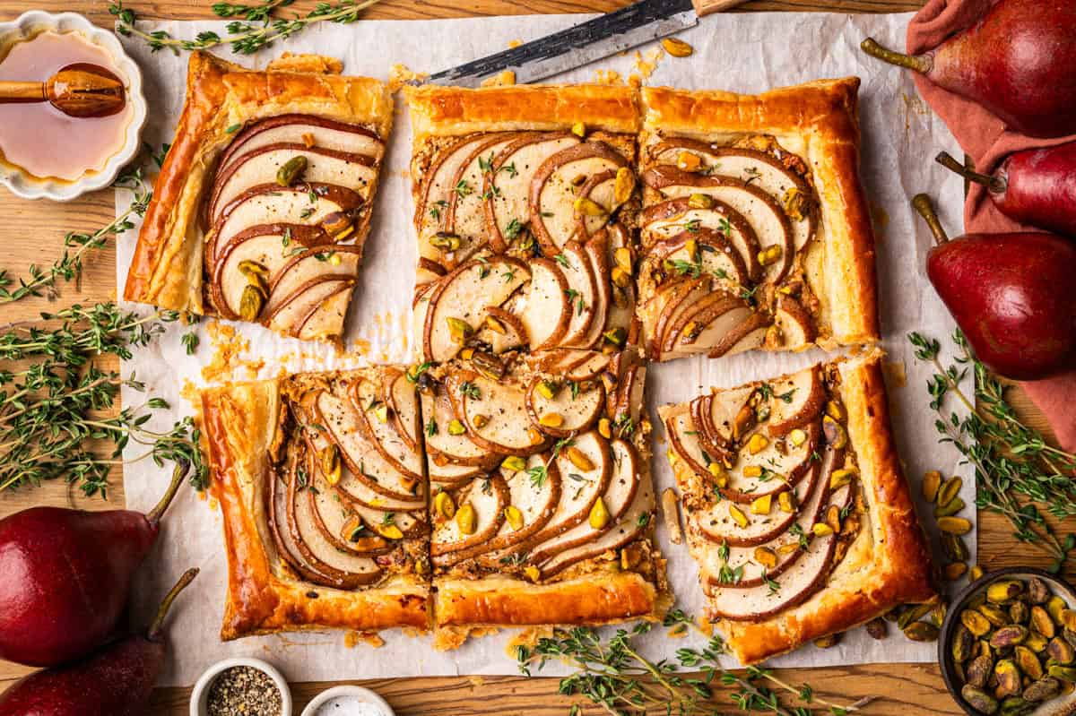 Pear Tart A rectangular pear tart with puff pastry, sliced pears, pistachios, and herbs, cut into pieces on parchment paper.