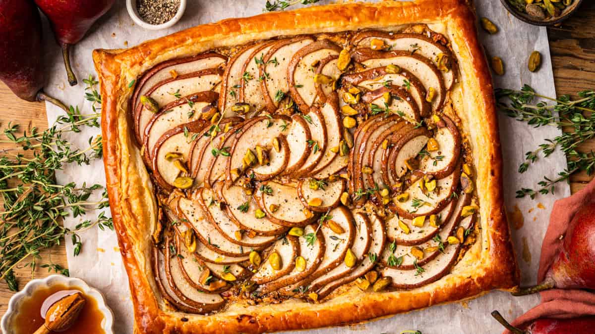 Pear Tart A rectangular pear tart with golden puff pastry, thinly sliced pears, chopped pistachios, and herbs on top, displayed on parchment paper.