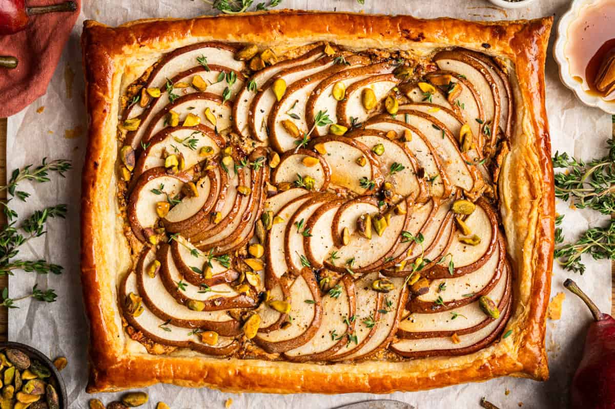 Pear Tart Golden-brown puff pastry tart topped with thinly sliced pears, chopped pistachios, and fresh thyme, arranged in a decorative pattern on parchment paper.