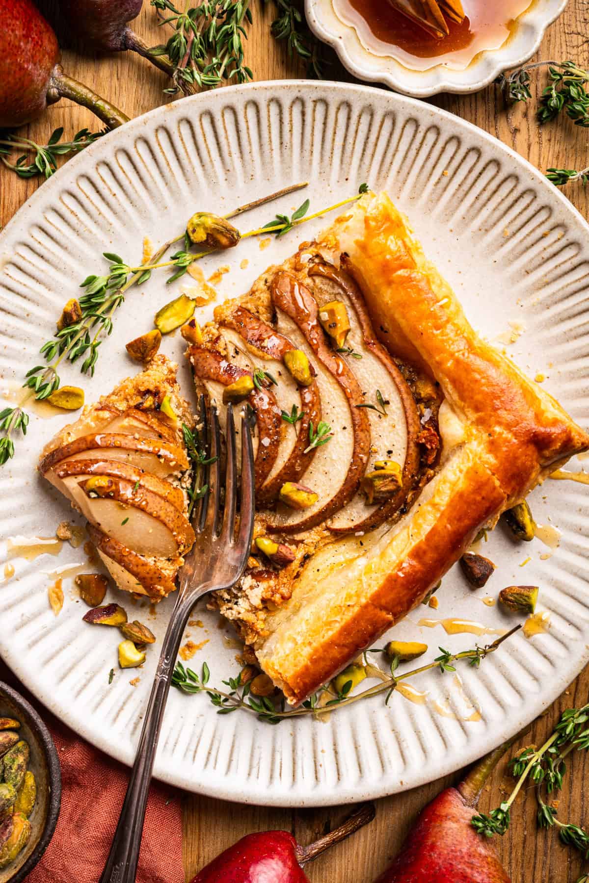 Pear Tart A slice of pear tart with golden crust, chopped pistachios, and thyme on a white plate.
