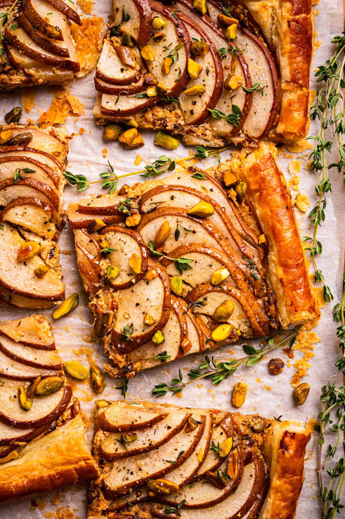 Pear Tart Rectangular slices of pear tart with puff pastry, topped with sliced pears, chopped pistachios, herbs, and a golden glaze, arranged on parchment paper.