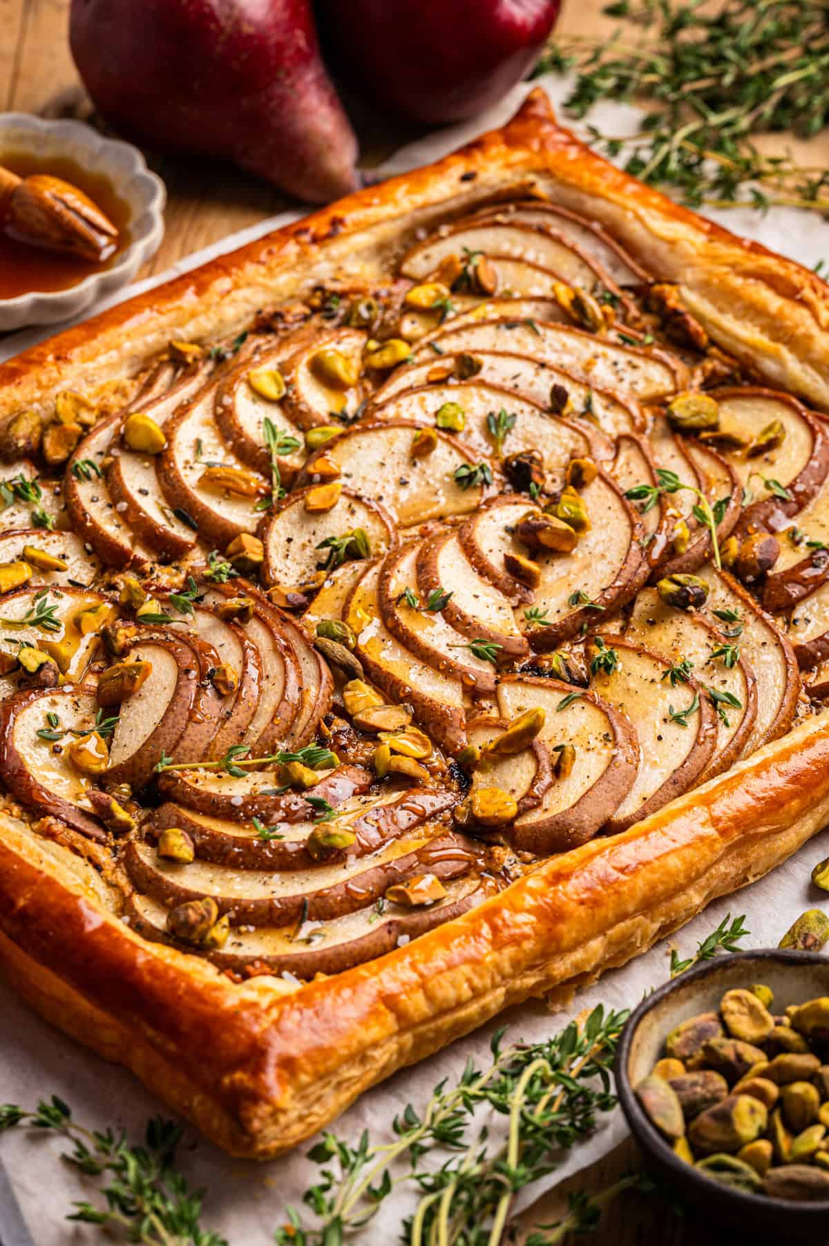 Pear Tart A rectangular puff pastry tart topped with thinly sliced pears, chopped pistachios, and fresh thyme, displayed on parchment paper.