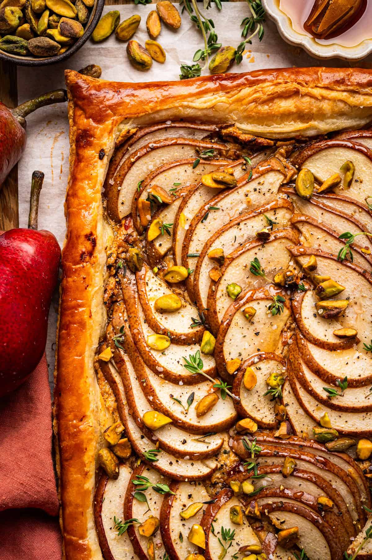 Pear Tart A golden-brown pear tart with sliced pears arranged in a spiral, topped with chopped pistachios and fresh herbs.