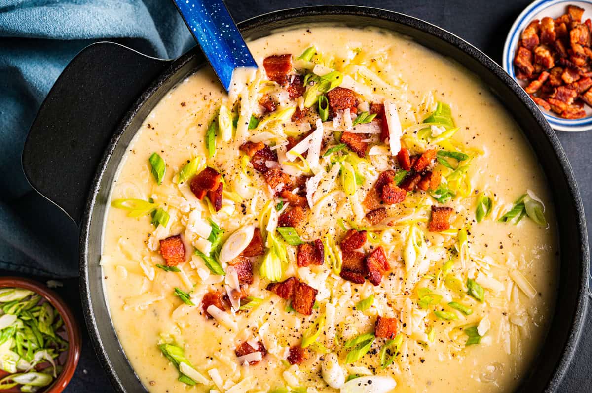 Creamy Potato Soup with Bacon and Cheddar A pot of creamy potato soup topped with chopped green onions, shredded cheese, crispy bacon pieces, and black pepper, with a blue ladle resting inside.