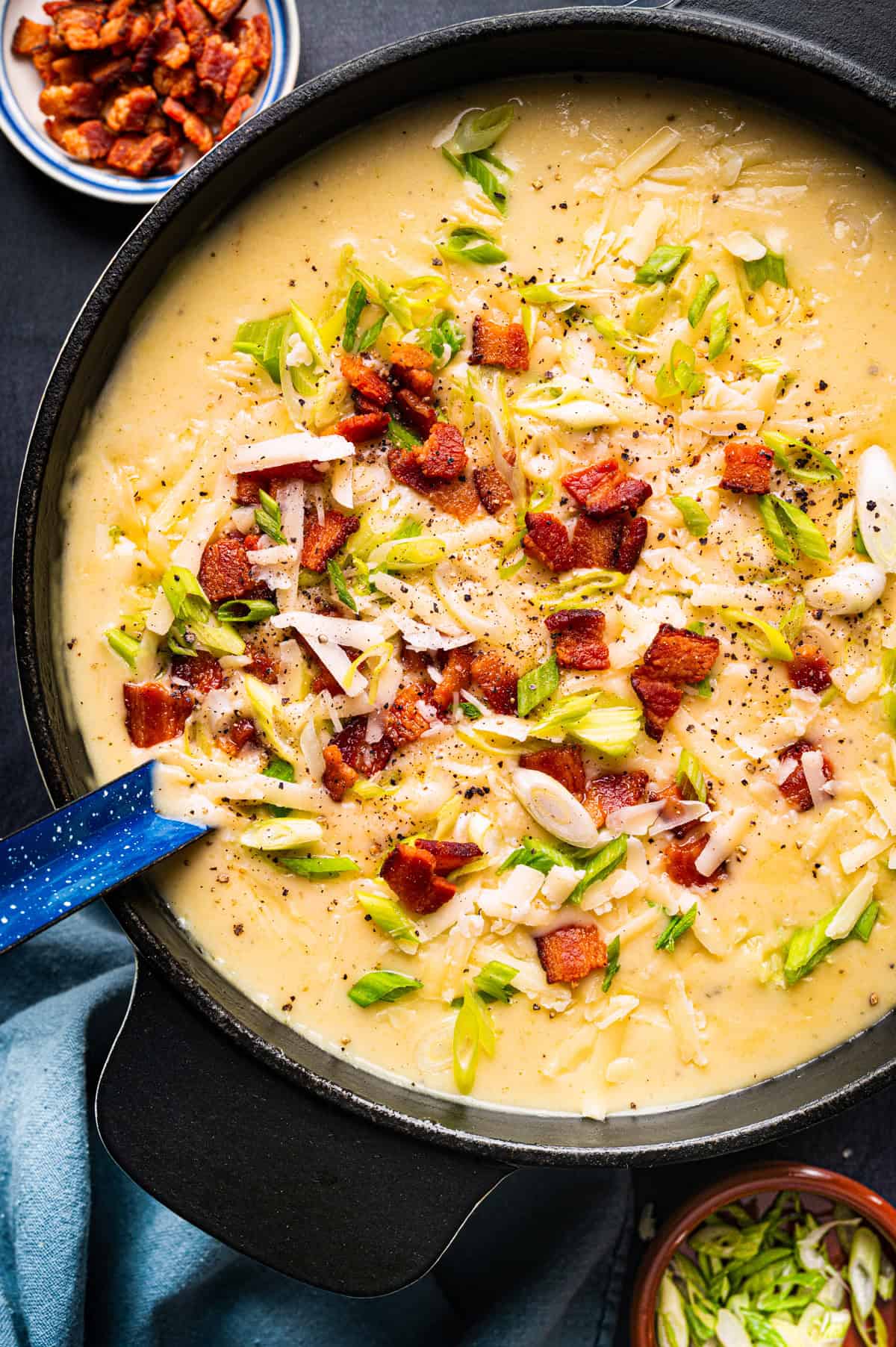 A pot of creamy potato soup topped with chopped bacon, shredded cheese, and sliced green onions, with a blue spoon resting inside. 