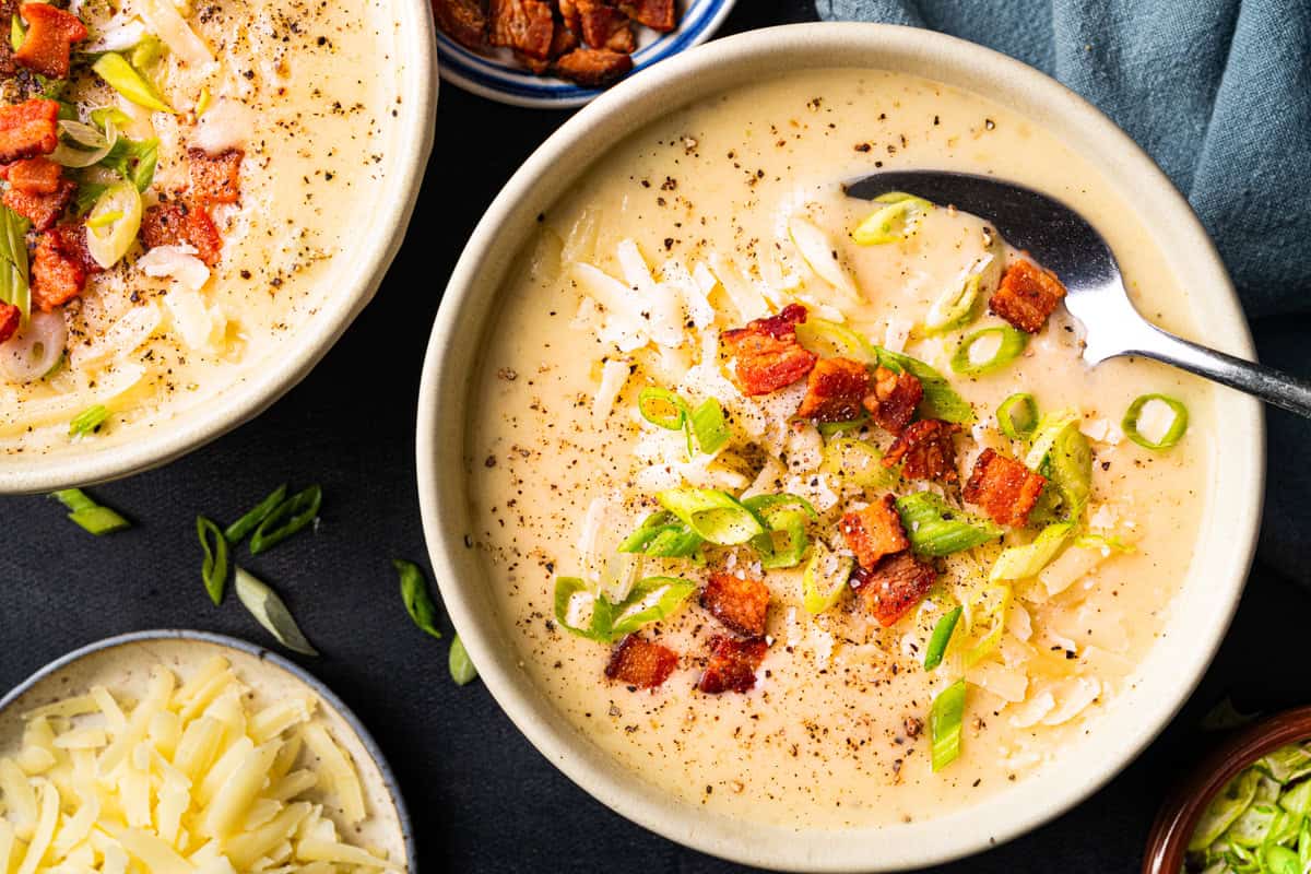 Bowl of creamy potato soup topped with shredded cheese, chopped green onions, bacon pieces, and black pepper, with a spoon inside.