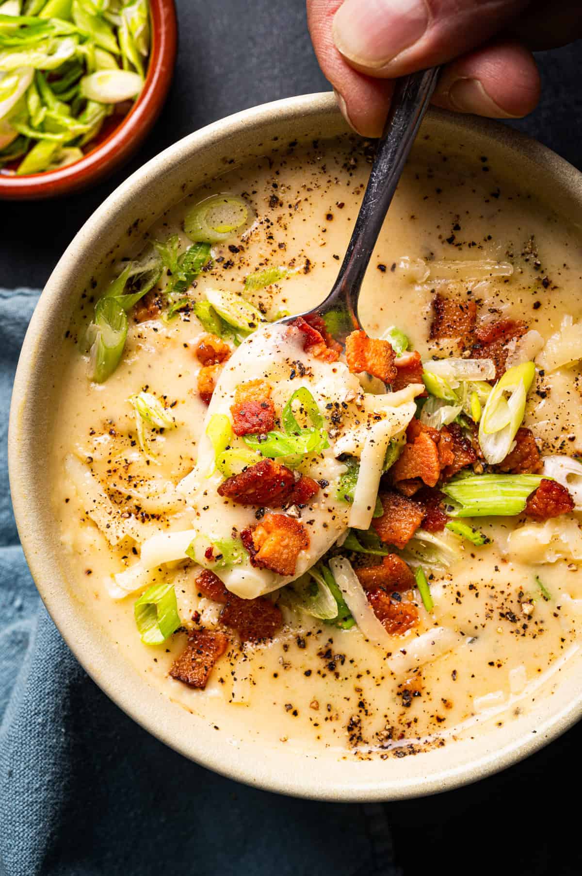 Creamy Potato Soup with Bacon and Cheddar A spoonful of creamy potato soup topped with crispy bacon, sliced green onions, shredded cheese, and black pepper, held above a bowl.