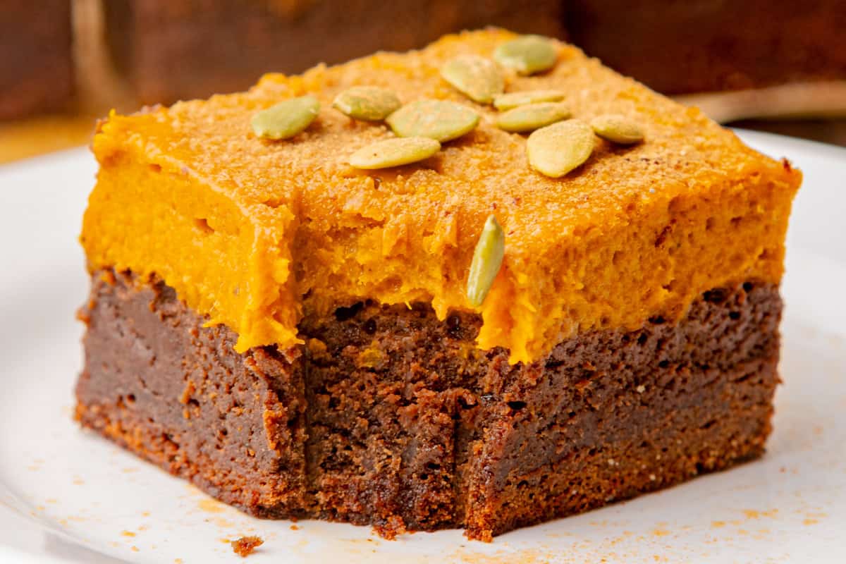 A close-up of a square dessert (pumpkin brownie) with dark chocolate base, a thick orange pumpkin layer on top, and garnished with pumpkin seeds. 