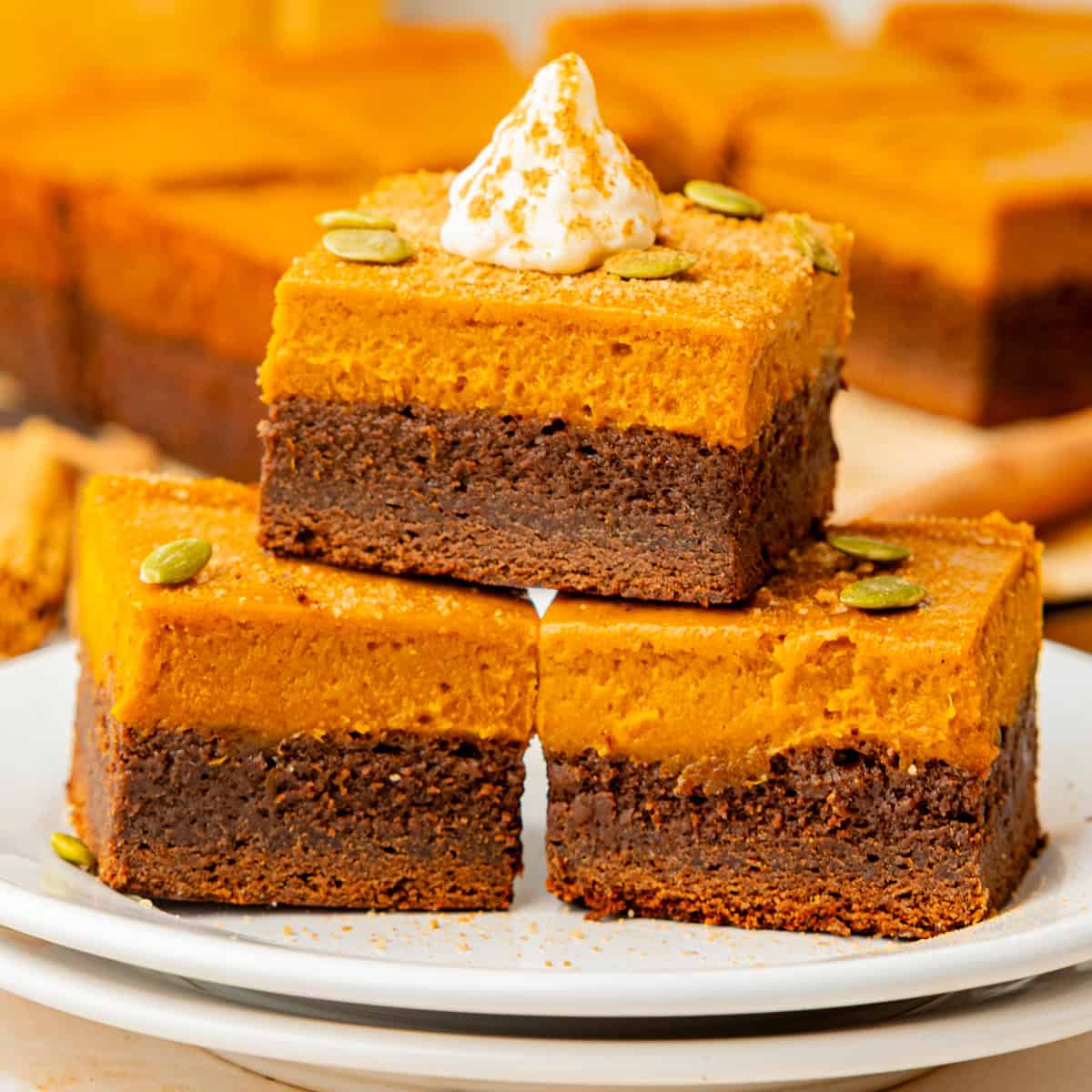 Pumpkin Brownies Three pumpkin chocolate brownies are stacked on a white plate. Each brownie has a chocolate base and a pumpkin layer, topped with whipped cream and pumpkin seeds.
