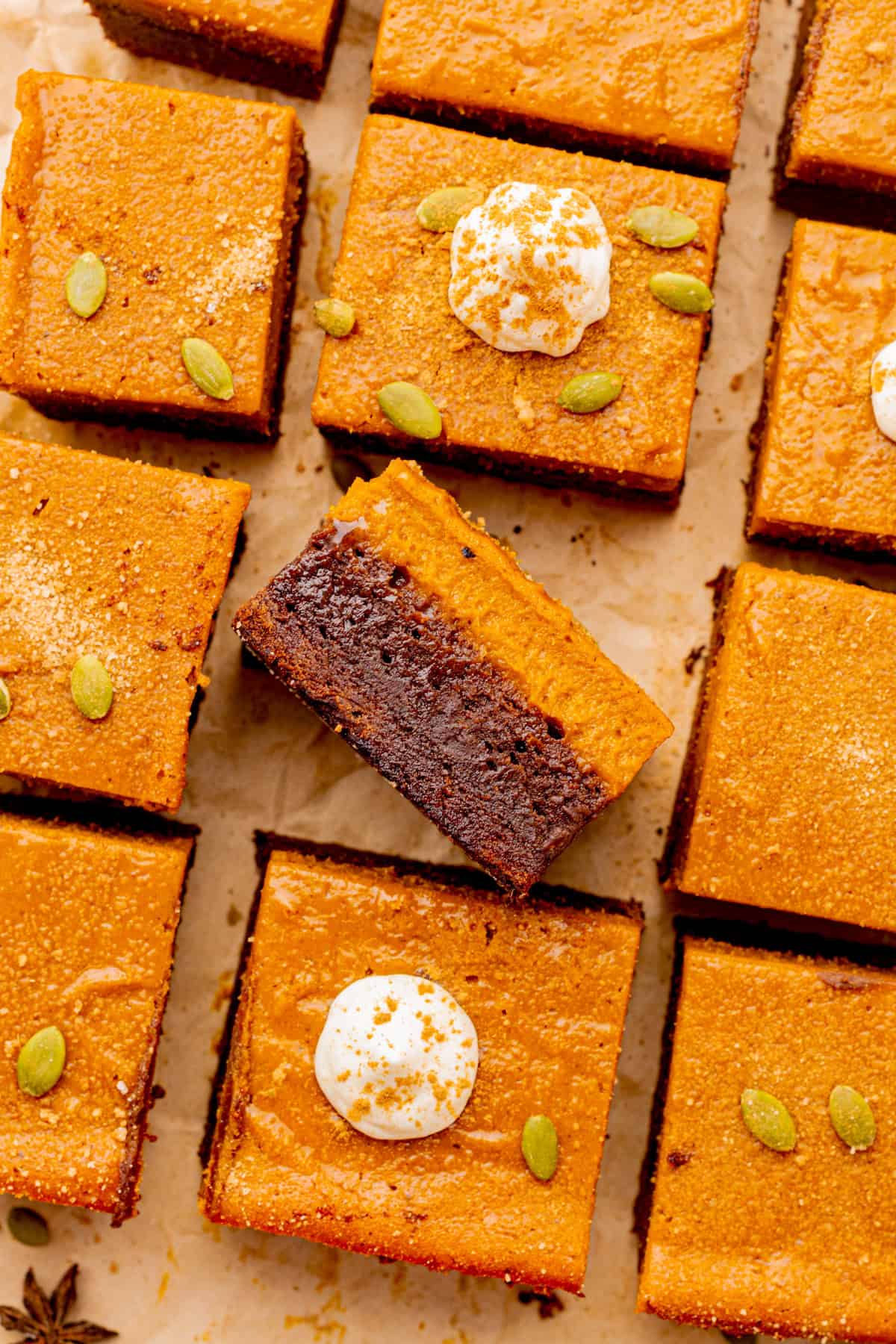 Pumpkin Brownies Pumpkin chocolate brownies cut into squares, some topped with whipped cream and pumpkin seeds. One brownie is turned on its side to show layers.