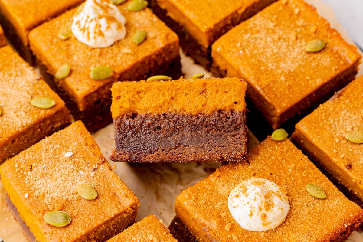 Pumpkin Brownies Pumpkin cheesecake brownies cut into squares, topped with whipped cream and pumpkin seeds. One brownie is lifted.