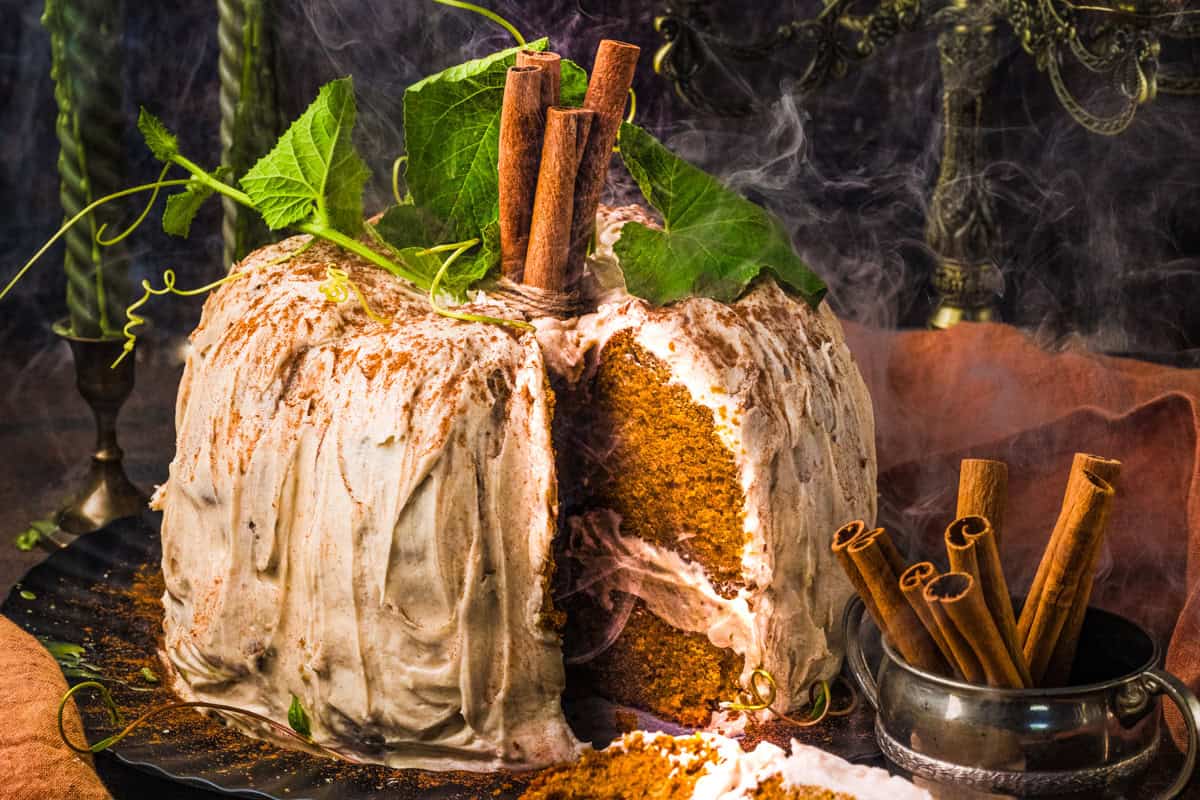 Pumpkin Shaped Cake for Halloween and Thanksgiving A spiced pumpkin cake with cream frosting is topped with cinnamon sticks and green leaves. One slice is cut out, revealing the moist interior.