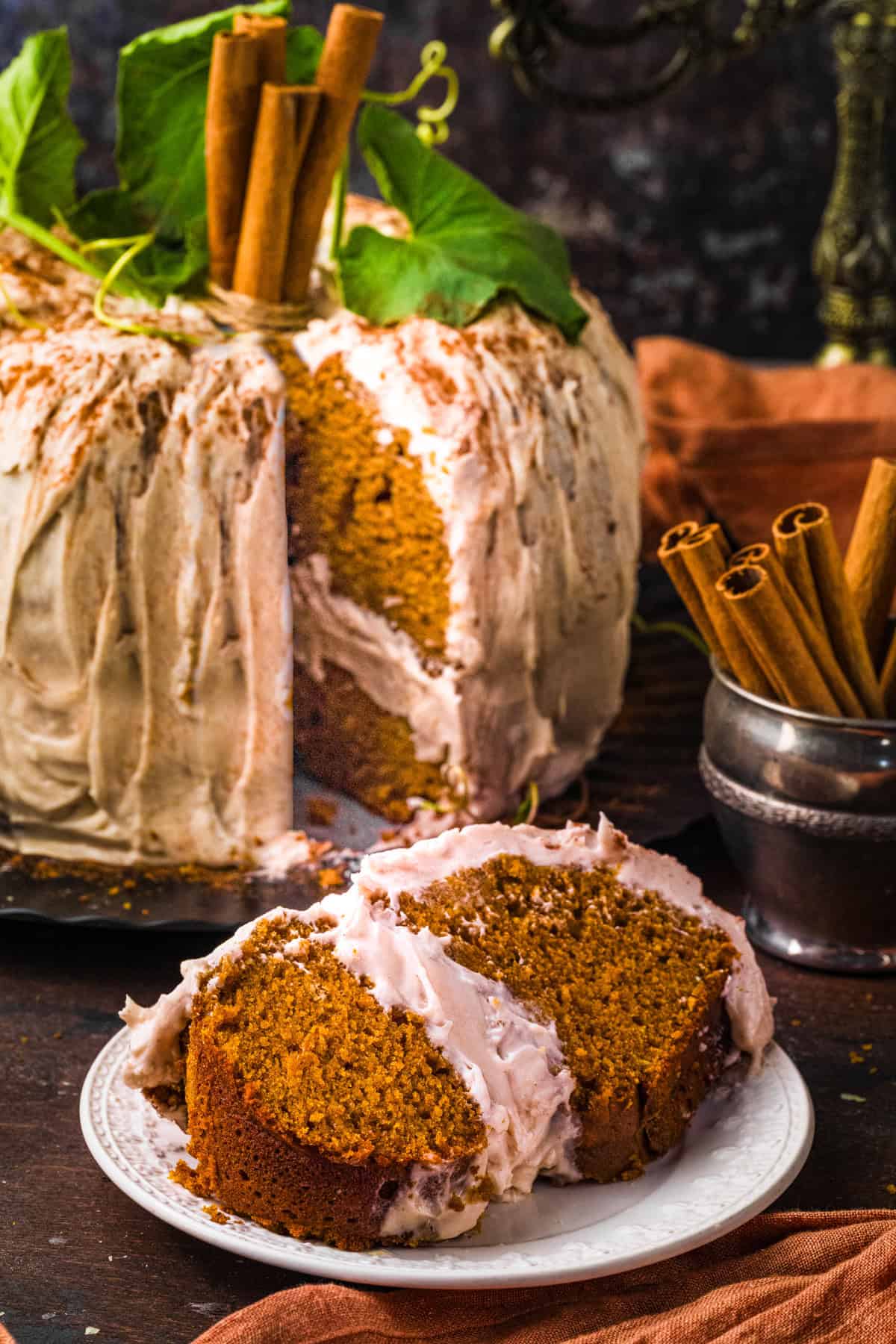 Pumpkin Shaped Cake for Halloween and Thanksgiving A frosted pumpkin cake with cinnamon sticks and green leaves sits on a table; A slice is served on a white plate.
