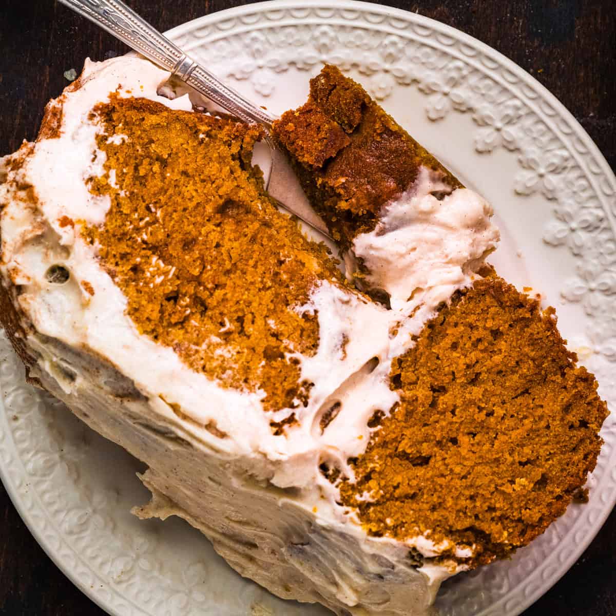 Pumpkin Cake for Halloween and Thanksgiving A close-up of a slice of spiced pumpkin cake with white frosting on a white plate.