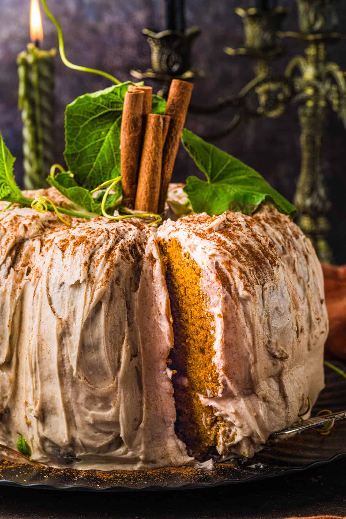 Pumpkin Shaped Cake for Halloween and Thanksgiving A pumpkin-shaped cake with cinnamon frosting, topped with green leaves and cinnamon sticks. A slice has been cut out, showing the golden interior.