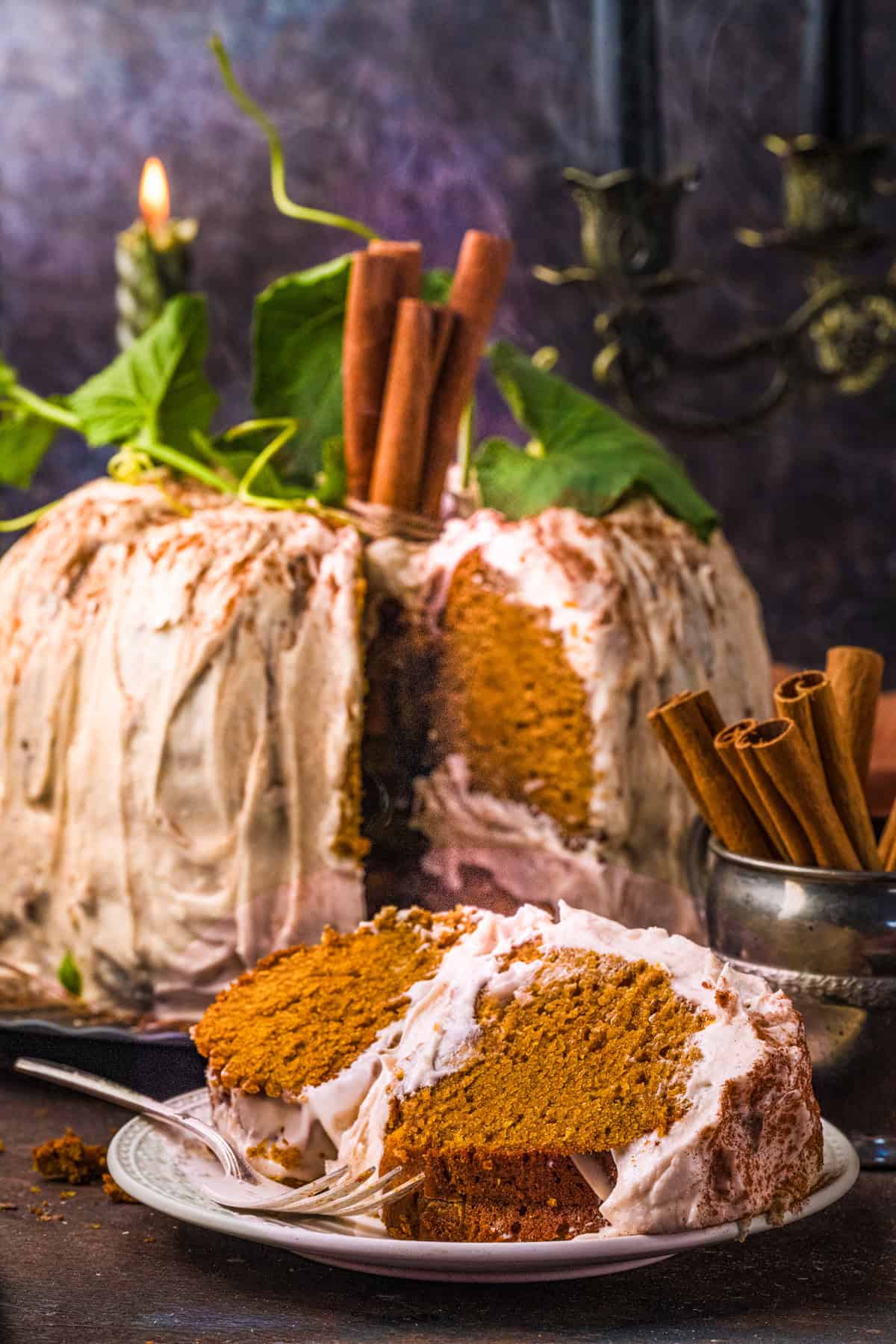 Pumpkin Shaped Cake with Cinnamon Butter Cream for Halloween and Thanksgiving A frosted pumpkin-shaped cake with cream cheese buttercream and cinnamon sticks and leaves on top sits on a table. A slice is served on a white plate in front.