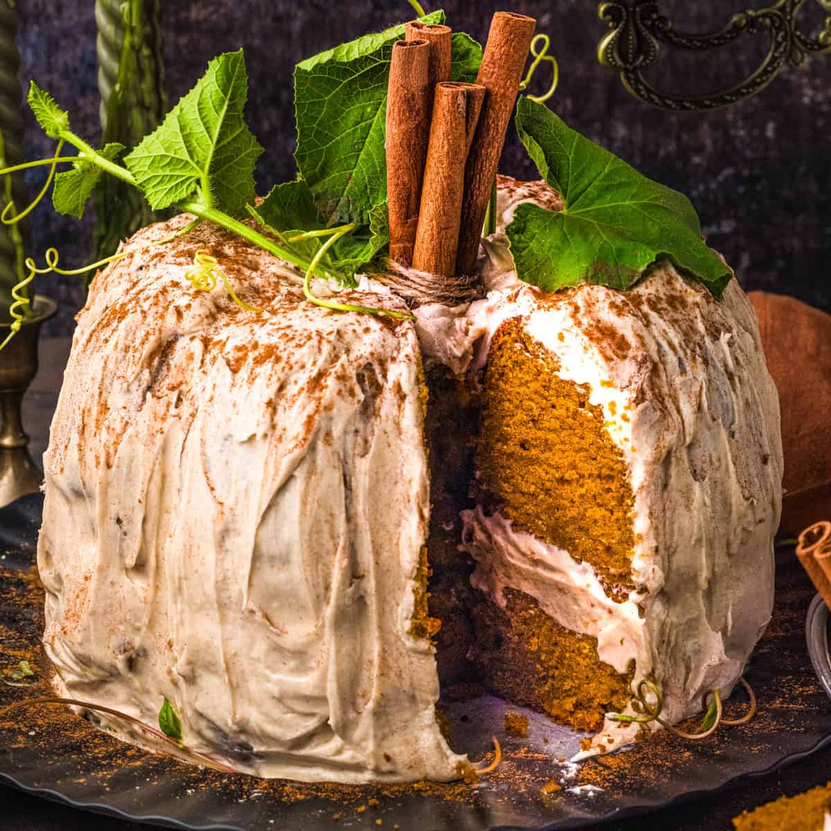 Pumpkin Shaped Cake for Halloween and Thanksgiving A frosted pumpkin-shaped bundt cake with a slice removed, topped with cinnamon sticks and green leaves, sits on a dark plate.