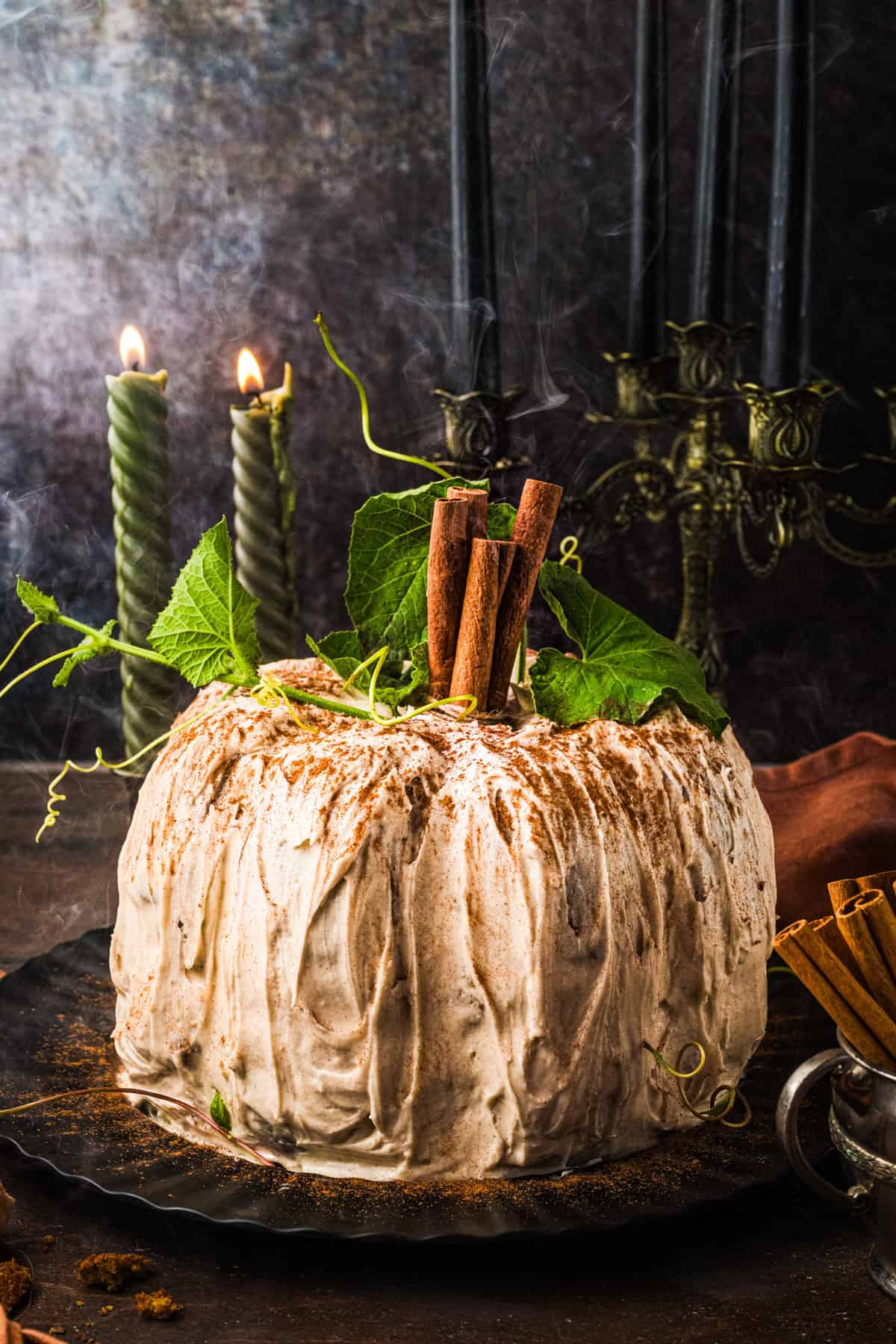 Pumpkin Shaped Cake for Halloween and Thanksgiving A frosted pumpkin-shaped cake topped with cinnamon sticks and green leaves sits on a dark plate.