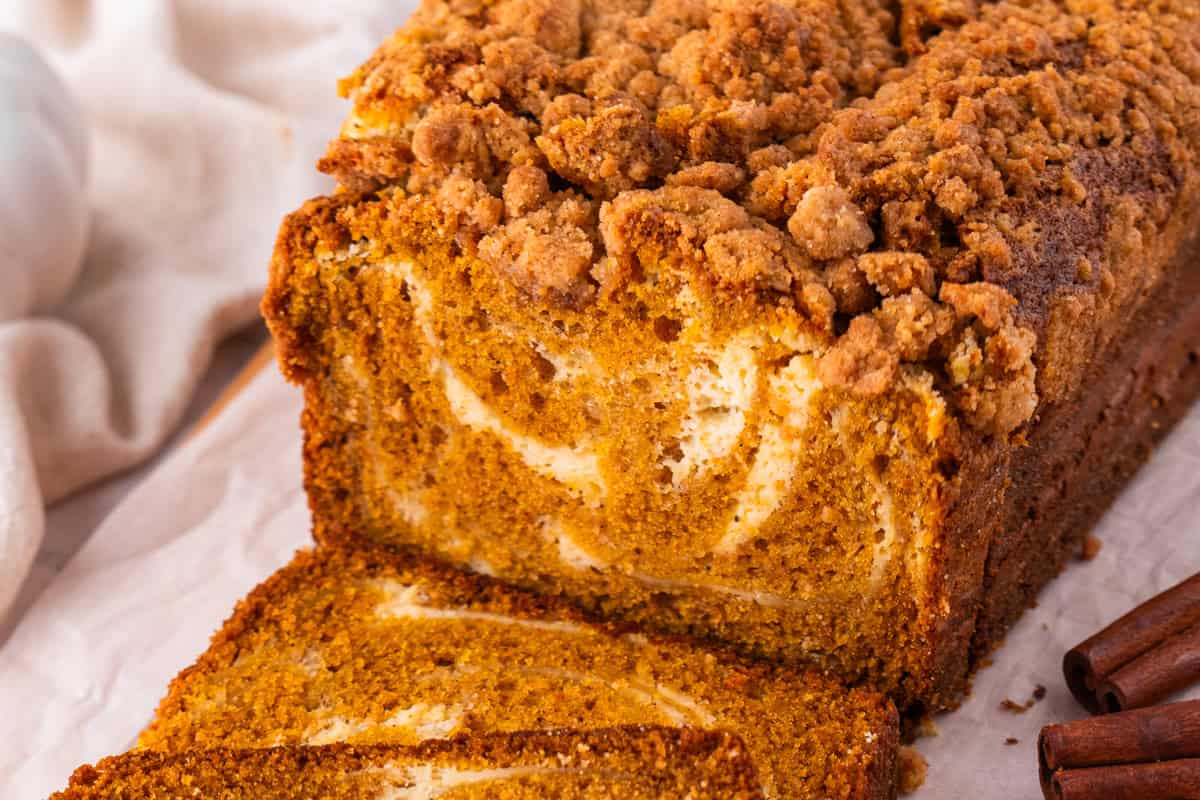 Pumpkin Cream Cheese Bread A loaf of pumpkin bread with a crumb topping and visible cream cheese swirls, with a slice cut and lying in front.