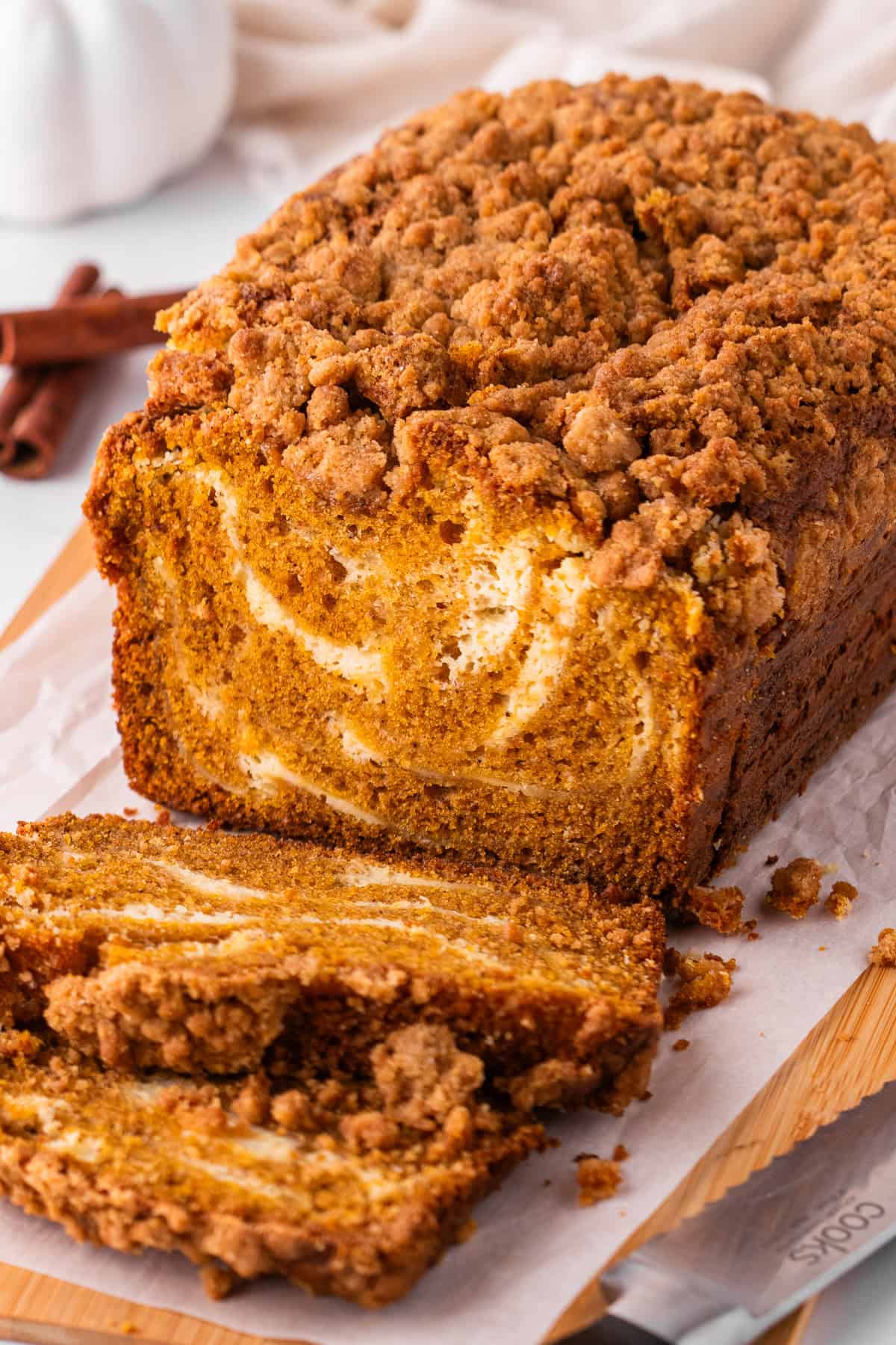 Pumpkin Cream Cheese Bread A loaf of pumpkin bread with a crumb topping and cream cheese swirl sits on parchment paper, with two slices cut in front.