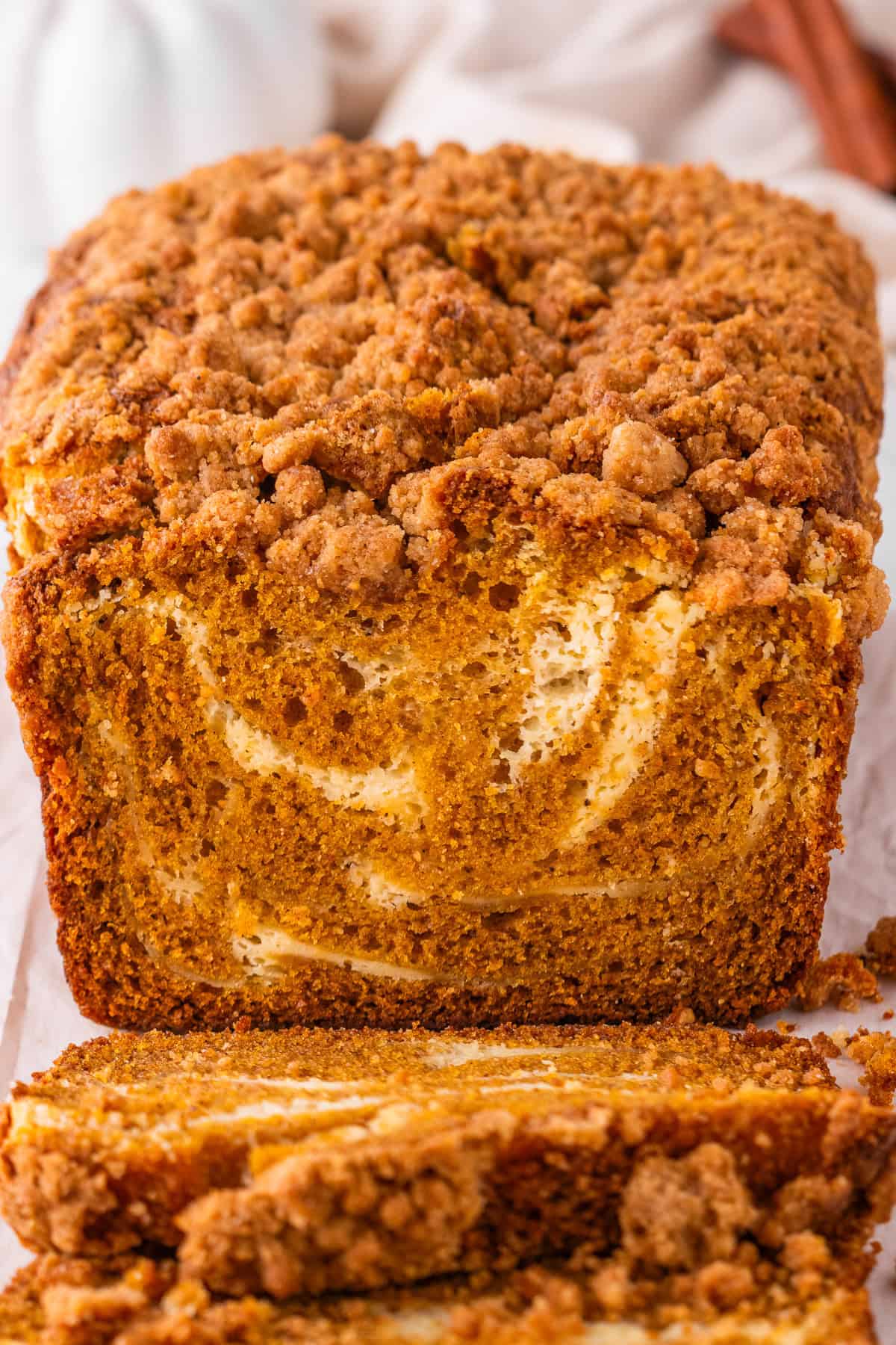 Pumpkin Cream Cheese Bread A close-up of a sliced loaf of pumpkin bread with a crumb topping and visible cream cheese swirl inside, resting on parchment paper.