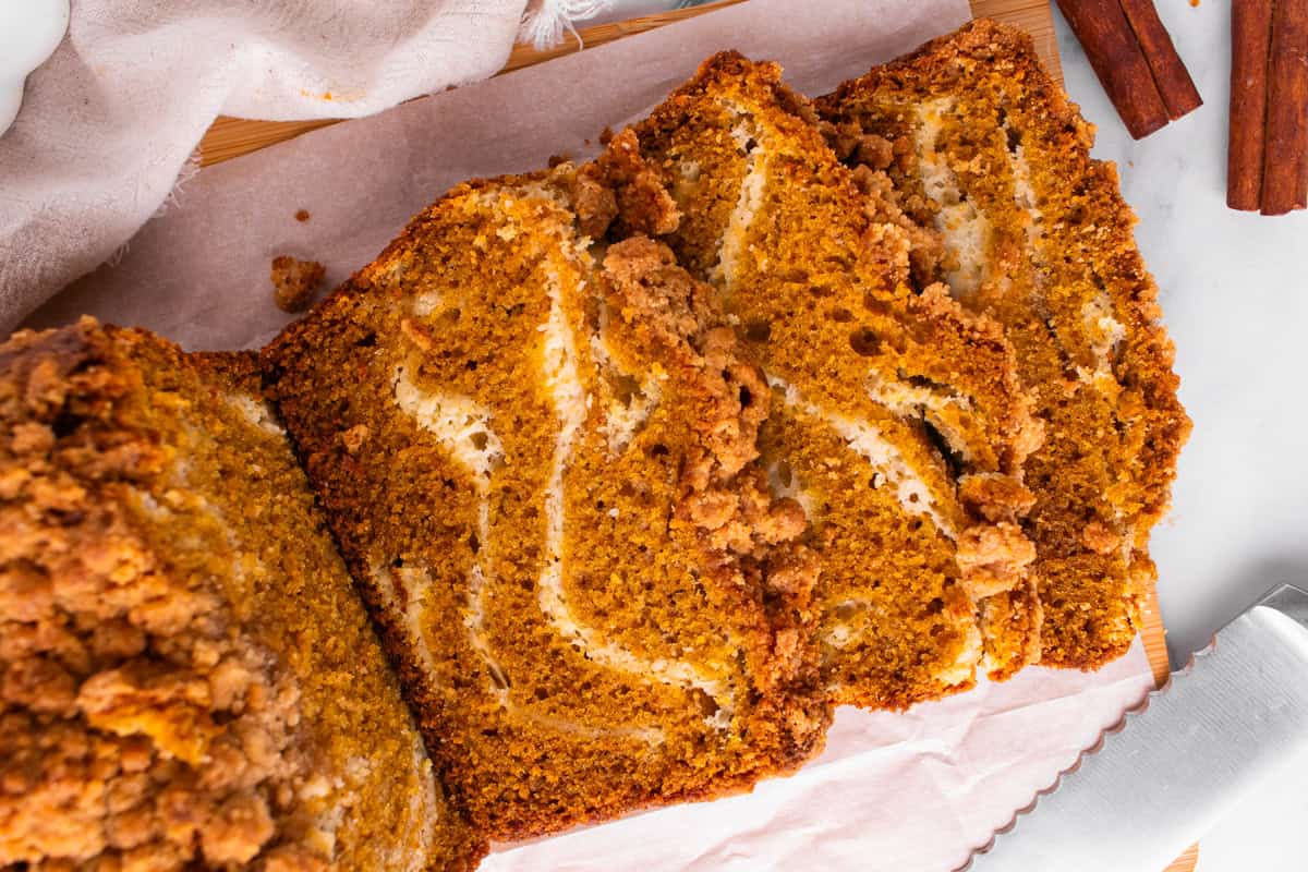 Pumpkin Cream Cheese Bread Three slices of pumpkin bread with a crumb topping and cream cheese swirl are arranged on parchment paper next to a loaf.