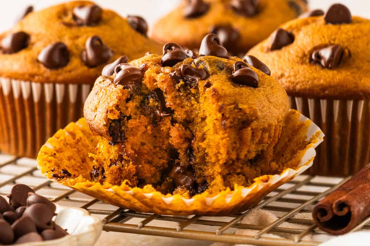 Pumpkin Chocolate Chip Muffins A close-up of a chocolate chip muffin with a bite taken out, resting in a paper liner on a cooling rack.