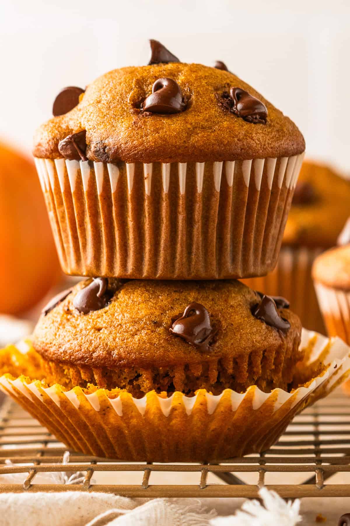 Pumpkin Chocolate Chip Muffins Two pumpkin chocolate chip muffins, one stacked on top of the other, sit on a wire rack. The bottom muffin has its paper liner partially peeled away, revealing its moist texture and chocolate chips.