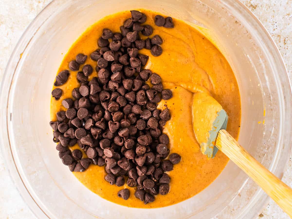 Pumpkin Chocolate Chip Muffins A glass bowl with pumpkin-colored batter, lots of chocolate chips on top, and a spatula resting inside, ready for mixing.