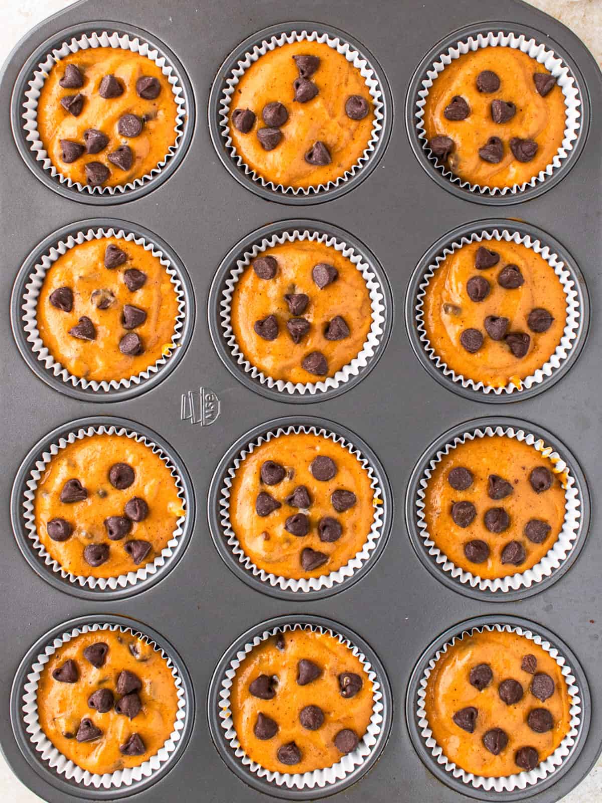 Pumpkin Chocolate Chip Muffins A muffin tin filled with twelve paper liners, each containing unbaked muffin batter topped with chocolate chips, ready to be baked.