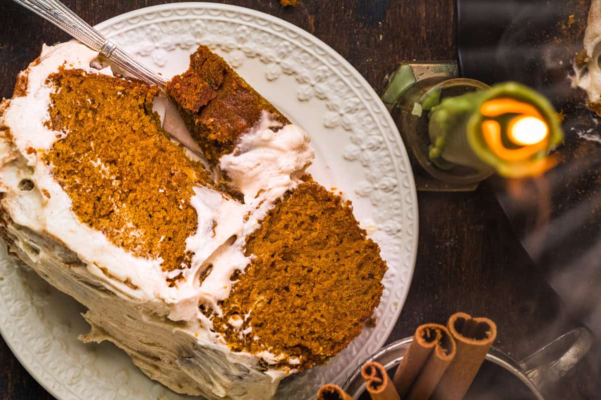 Pumpkin Shaped Cake with Cinnamon Buttercream Frosting for Halloween and Thanksgiving A slice of spiced pumpkin cake with cream frosting sits on a white plate with a fork. Nearby are a lit candle, a glass, and some cinnamon sticks on a dark wooden table.