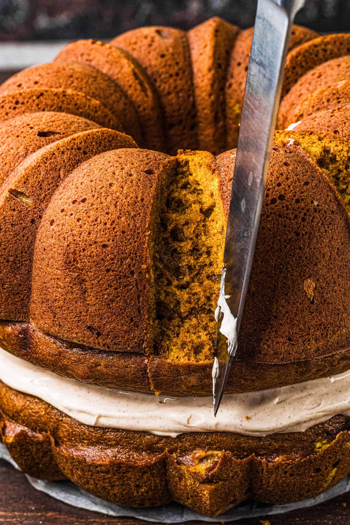 Pumpkin Shaped Cake with Cinnamon Butter Cream for Halloween and Thanksgiving A knife slices into a round bundt cake with a creamy filling layer in the middle, revealing its moist, golden interior.
