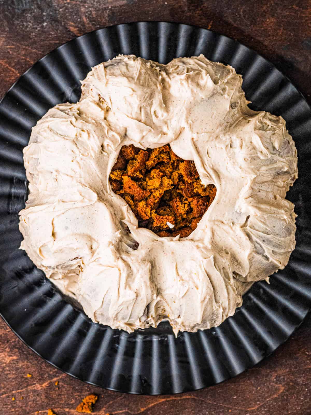 Pumpkin Shaped Cake with Cinnamon Buttercream Frosting for Halloween and Thanksgiving A round pumpkin cake with light brown frosting on a black plate, with crumbled cake pieces in the center.