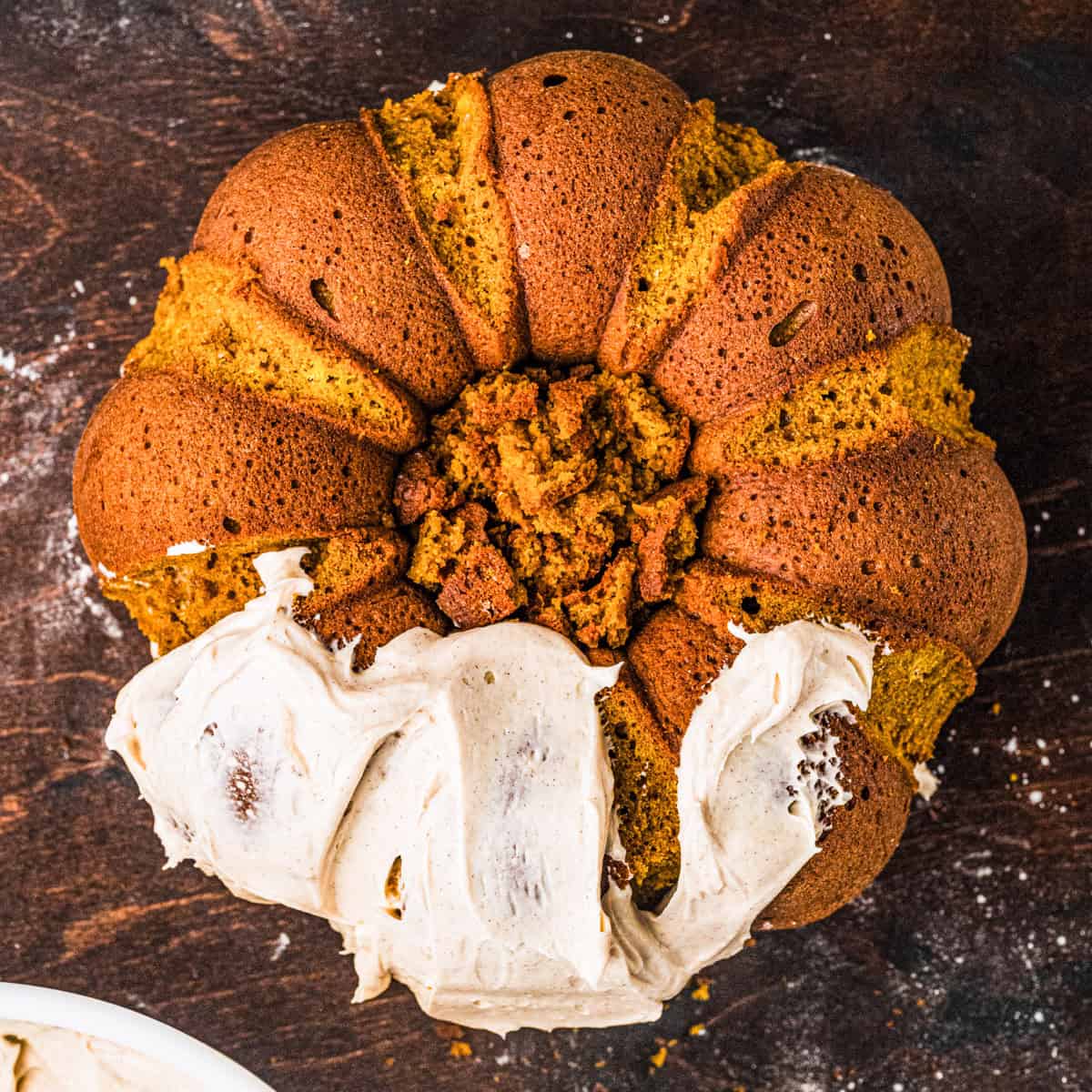 Pumpkin Shaped Cake with Cinnamon Butter Cream for Halloween and Thanksgiving A pumpkin bundt cake with cinnamon frosting partially spread on top, sitting on a dark wooden surface, with some crumbs in the cakes center.