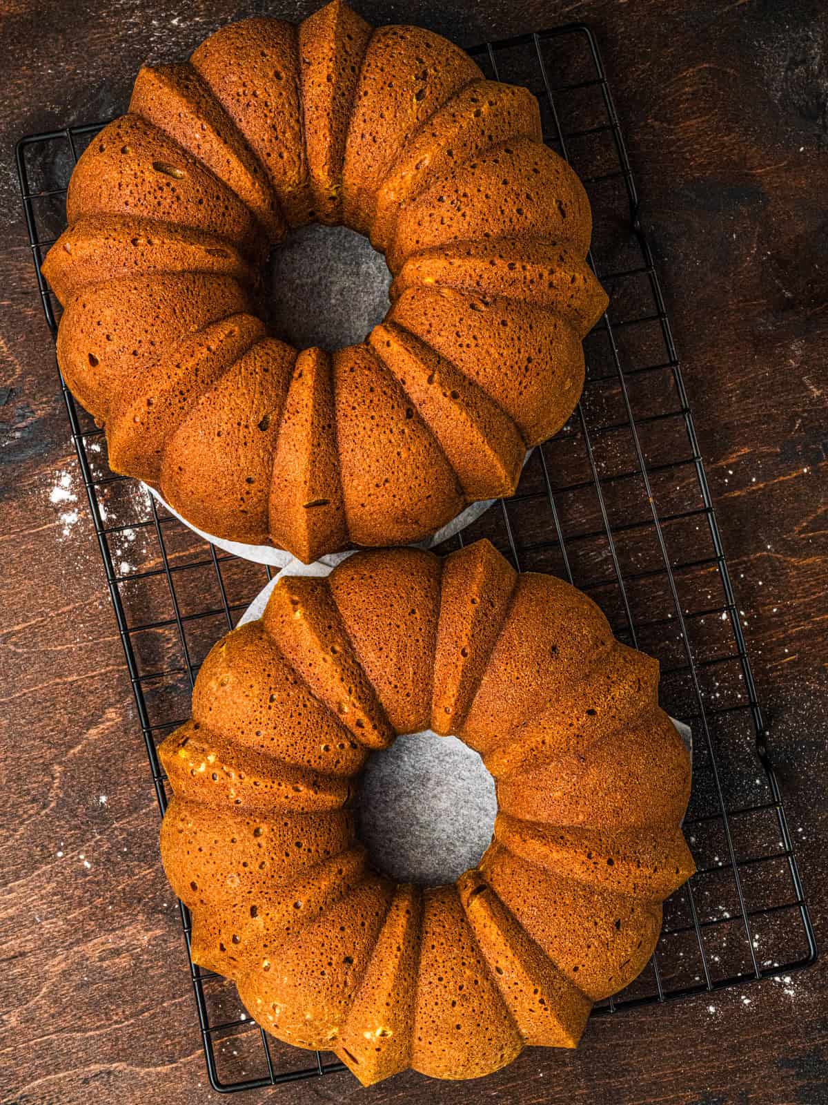 Pumpkin Bundt Cakes Two golden brown pumpkin bundt cakes with ridged edges cooling on a black wire rack atop a dark wooden surface.