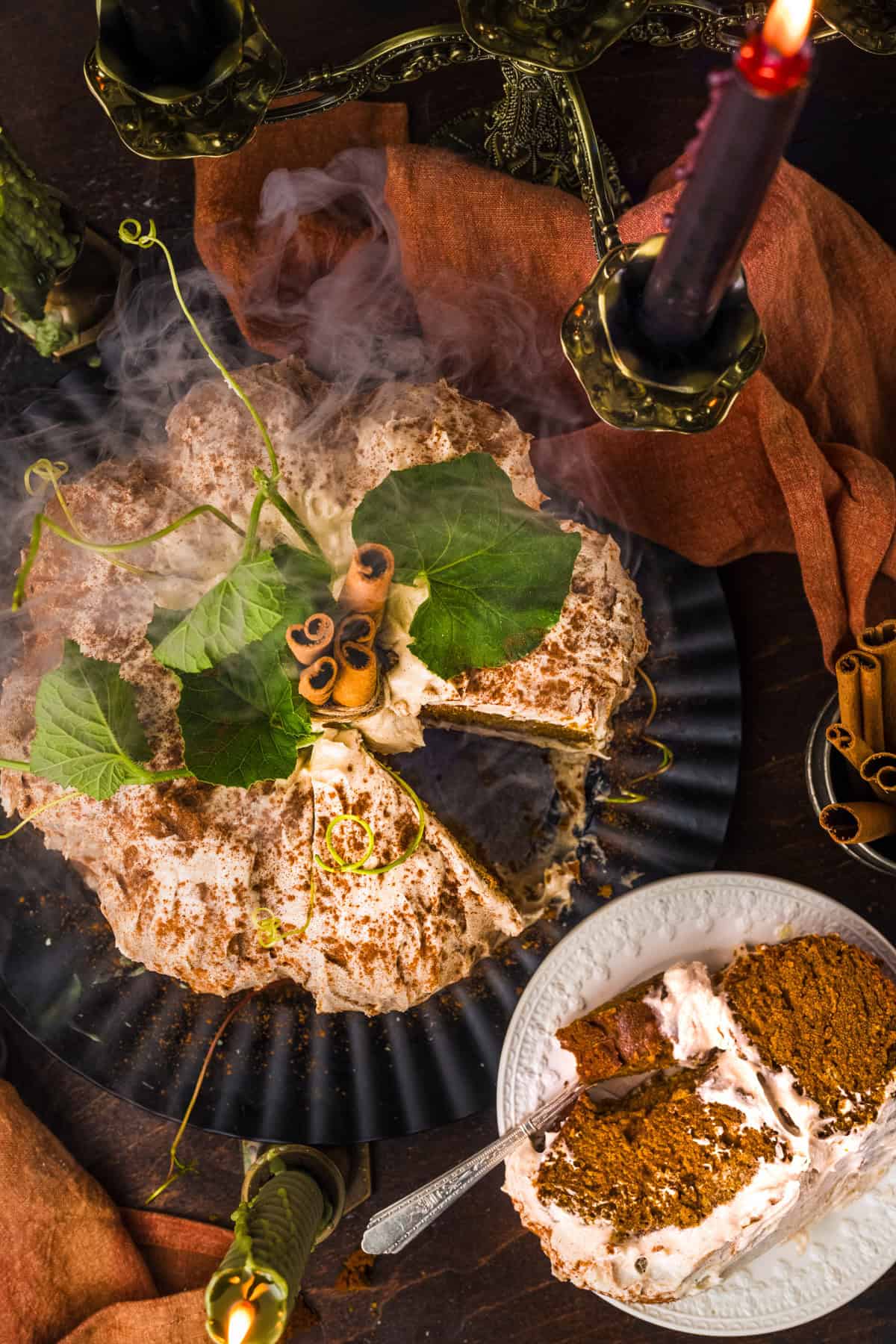 Pumpkin Shaped Cake for Halloween and Thanksgiving A sliced pumpkin-shaped cake topped with leaves, cinnamon sticks, and mist, on a dark plate. A piece is served on a white dish beside lit candles and a rust-colored cloth.