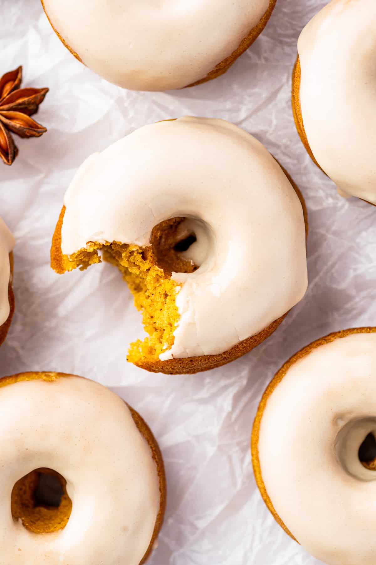 Pumpkin Spice Donuts Iced donuts on white parchment paper, one with a bite taken out. The donuts have light-colored glaze.