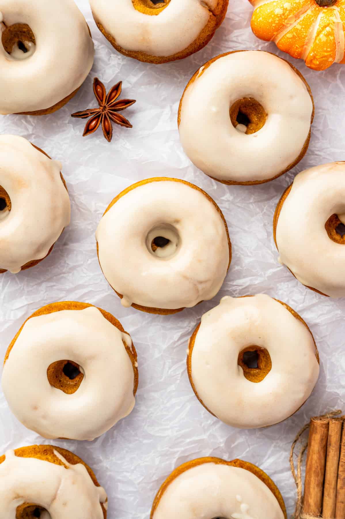 Pumpkin Spice Donuts Donuts are arranged on a crinkled white surface with a small decorative pumpkin, star anise, and cinnamon sticks nearby.