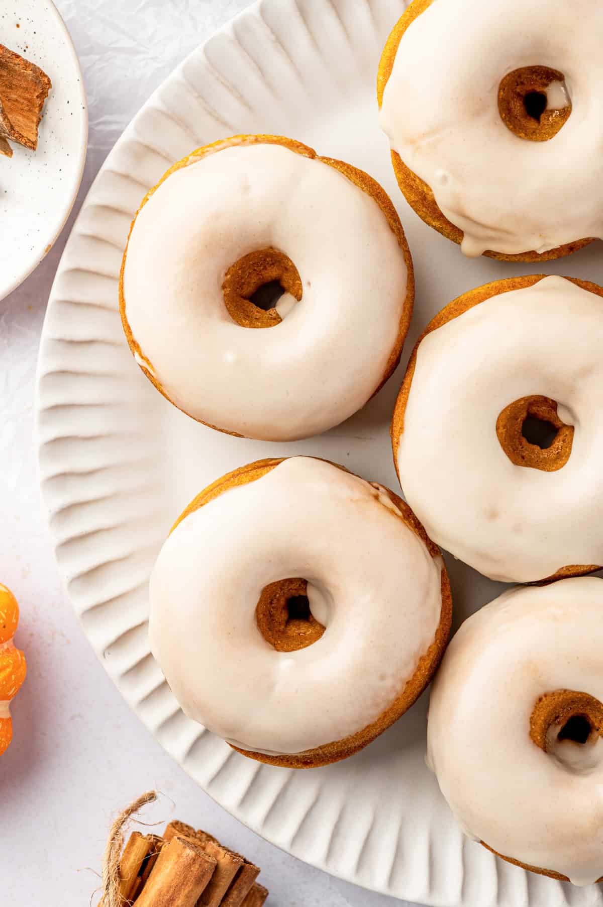 Pumpkin Spice Donuts Five glazed donuts are arranged on a white, textured plate. The donuts have a smooth, light icing and a golden-brown color.