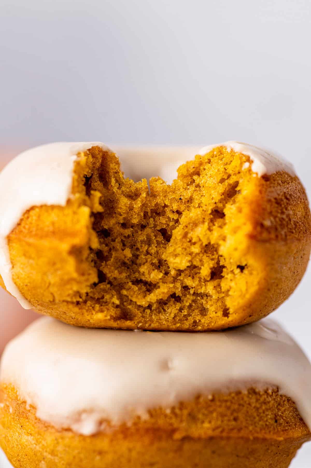 Pumpkin Spice Donuts A close-up of two iced pumpkin donuts stacked, with the top donut missing a bite, showing its moist orange interior and white glaze.