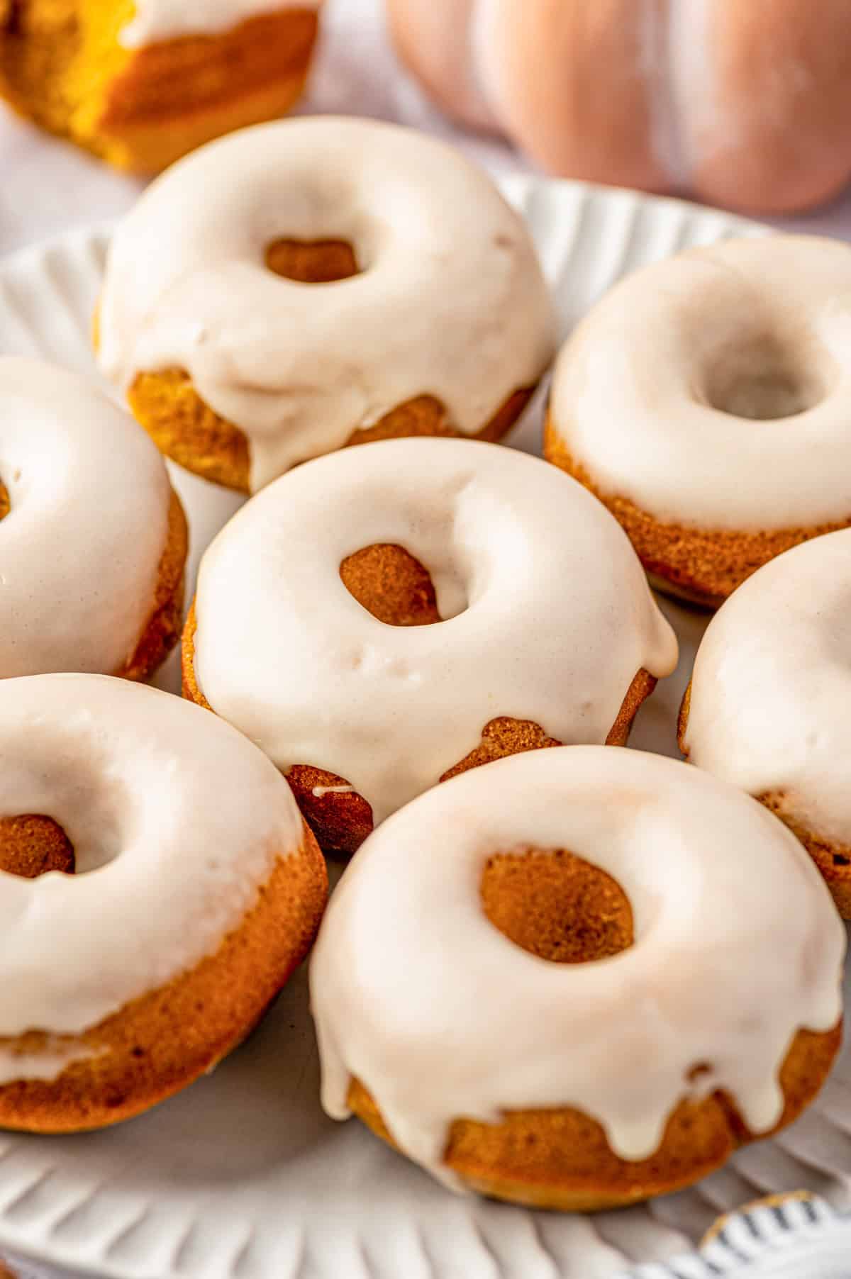 Pumpkin Spice Donuts A plate of baked donuts with light orange color, topped with smooth white glaze. The donuts have a soft texture and are arranged closely together on a white plate.