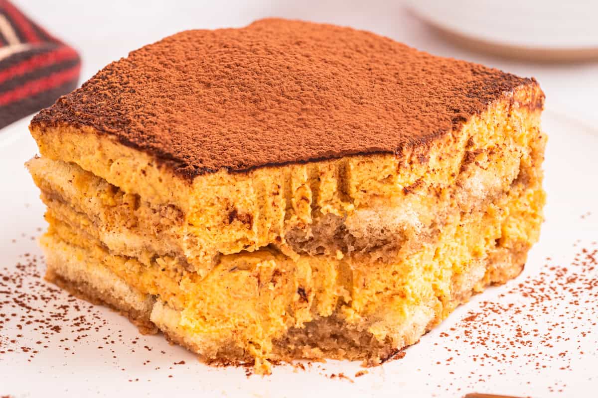 Pumpkin Tiramisu A close-up of a square slice of tiramisu dusted with cocoa powder, showing creamy layers of filling and soaked ladyfinger biscuits on a white plate.