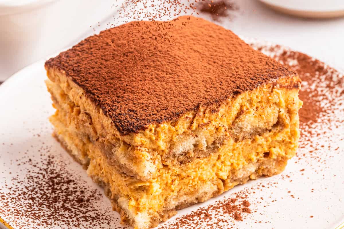 A close-up of a slice of tiramisu, showing creamy layers of mascarpone and coffee-soaked ladyfingers, dusted generously with cocoa powder on top and the plate.