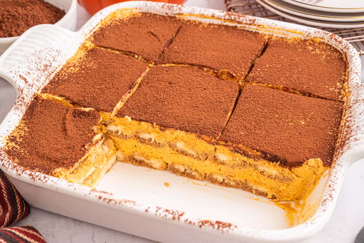 Pumpkin Tiramisu A baking dish with nine squares of tiramisu, dusted with cocoa powder. Several pieces are missing from the bottom right corner, revealing creamy layers and ladyfinger biscuits inside.