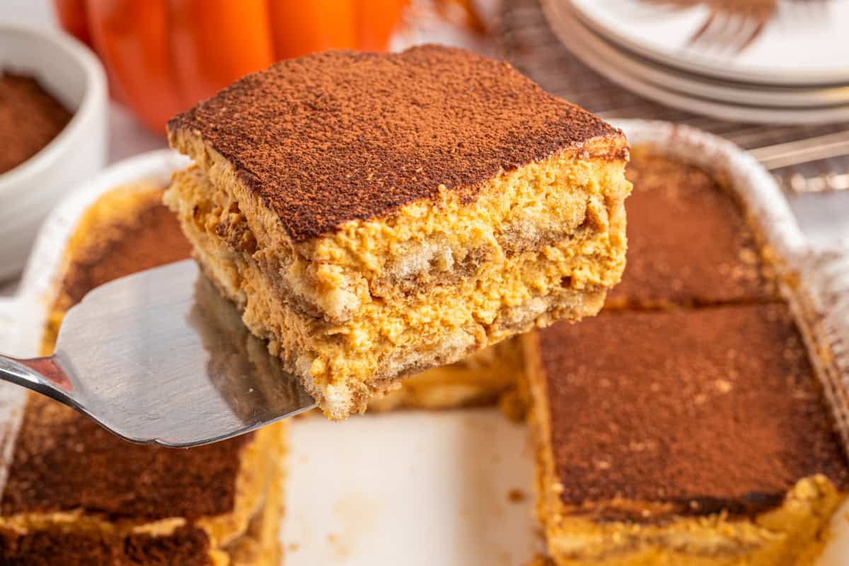 Pumpkin Tiramisu A close-up of a square slice of pumpkin tiramisu being lifted from a tray, showing creamy layers topped with cocoa powder.