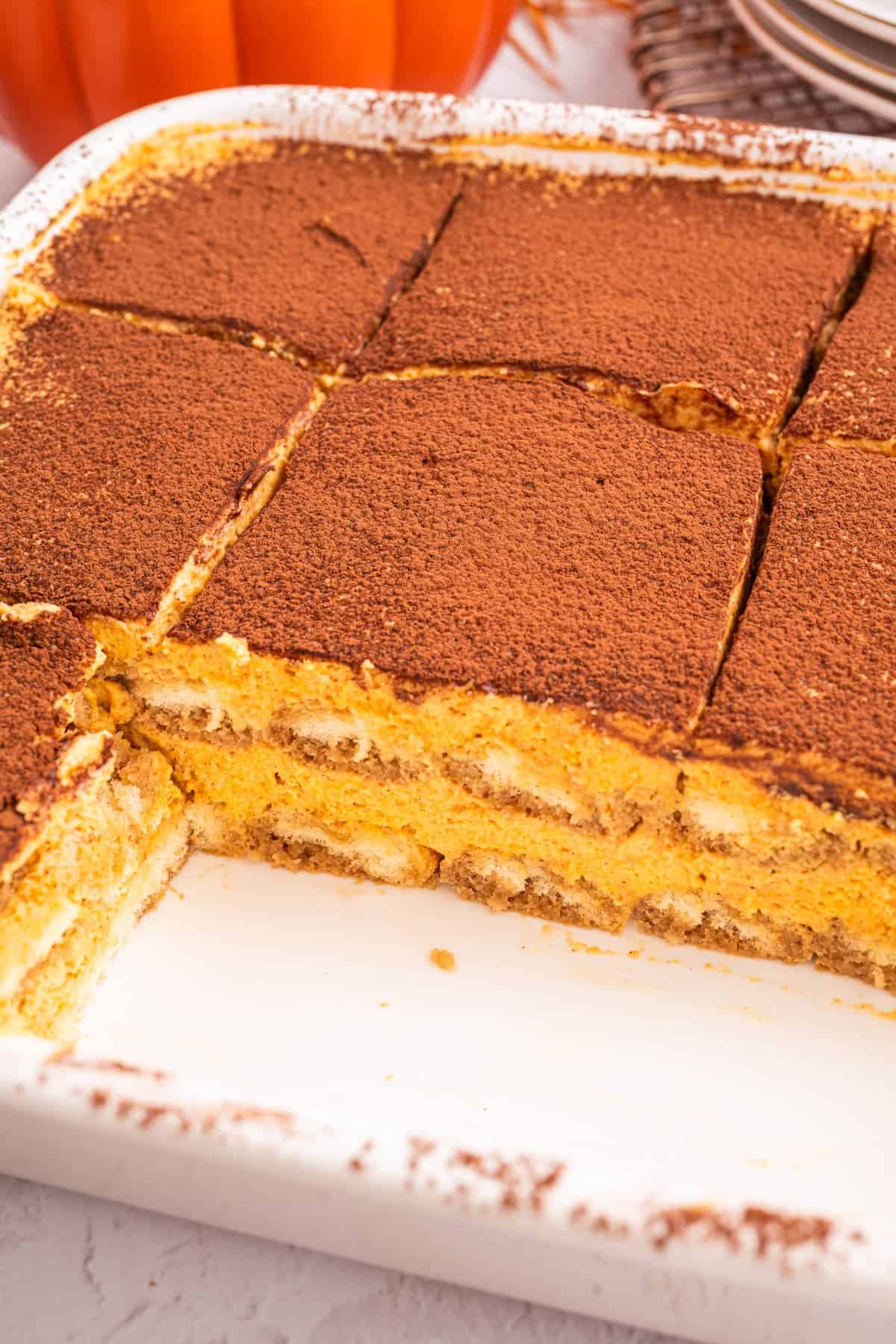 Pumpkin Tiramisu A rectangular dish of pumpkin tiramisu cut into squares, with a layer of cocoa powder on top and creamy pumpkin filling visible where a piece has been removed.